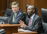 Representative Noor sits at the testifiers' table and speaks to someone out of view. Mike Opat sits next to him and watches as he speaks.